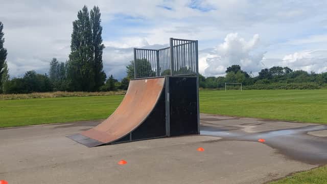 Skate ramp with a metal frame on a concrete surface, surrounded by grass and trees under a cloudy sky.