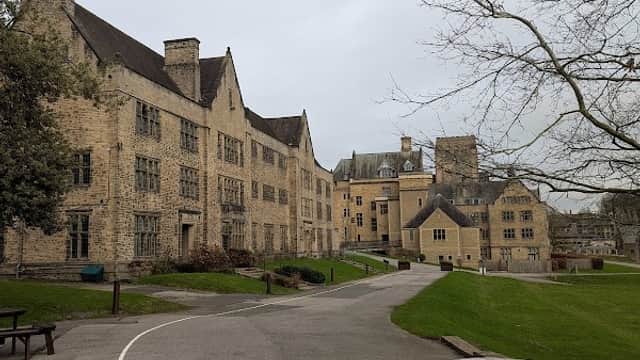 Stone buildings with large windows line a pathway, surrounded by grass and trees, under a cloudy sky.