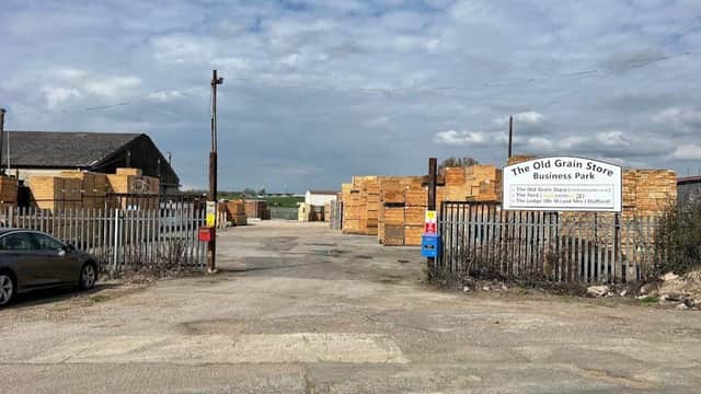 Entrance to The Old Grain Store Business Park, with stacked wooden crates and a gravel road under a cloudy sky.