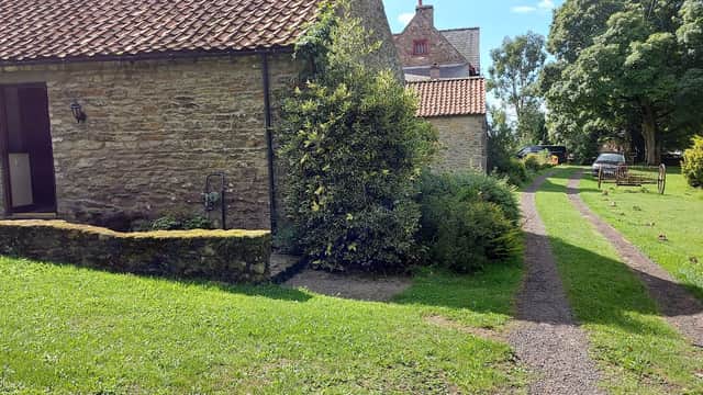 Stone cottage with a red-tiled roof, gravel path, lush greenery, and parked car in a tranquil Yorkshire setting.