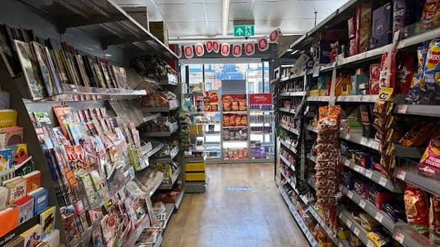 Aisle in a Co-op Food store with shelves stocked with magazines, snacks, and drinks, leading to the entrance.