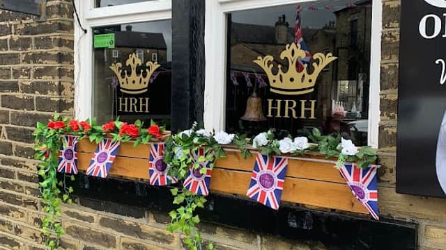 Bar windows adorned with flowers, Union Jack flags, and a crown logo, set against a stone wall in Wibsey, Yorkshire.