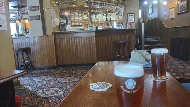 Two pints of dark beer on a wooden table, with a rustic bar and decorative lights in the background.