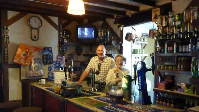 Bar interior featuring a counter, two people smiling, shelves of drinks, and a vintage clock on the wall.
