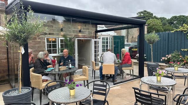 Glass-walled bistro with outdoor seating, guests enjoying drinks, and potted plants in a cozy setting.