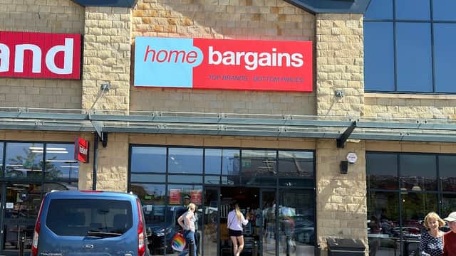 Storefront of Home Bargains with a large red sign, shoppers entering, and a blue vehicle parked outside.