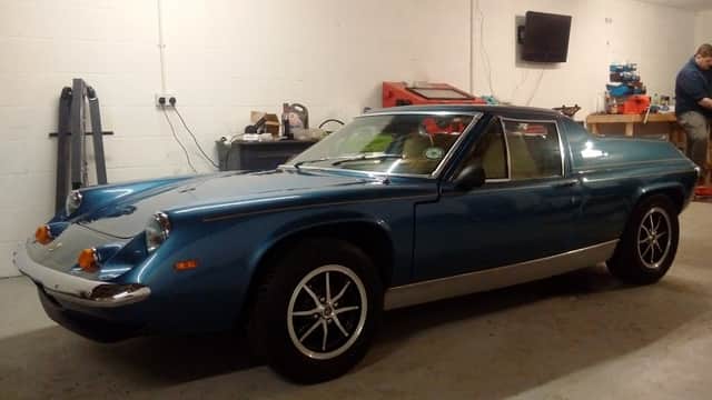 Classic blue sports car in a garage, with tools and equipment visible in the background.