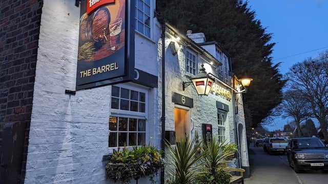 Quaint bar exterior with a sign reading "The Barrel," surrounded by greenery and lit by evening lights.