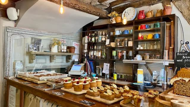 Cozy cafe interior with a wooden counter, baked goods, colorful mugs, and a rustic shelf filled with jars and bottles.