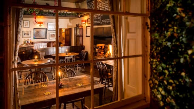 Cozy pub interior with wooden tables, chairs, a fireplace, and warm lighting, seen through a window.