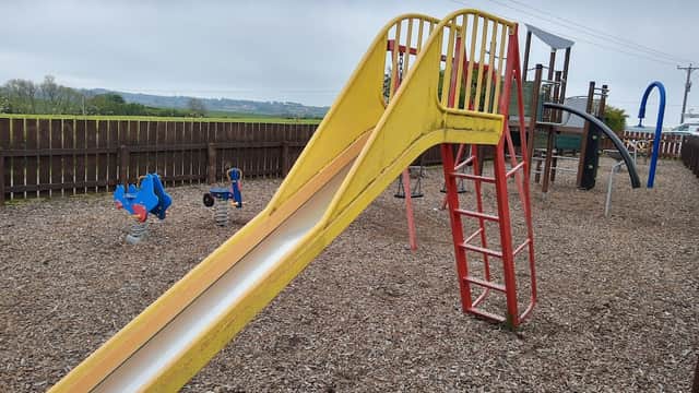 Bright yellow slide and climbing frame in a playground surrounded by wooden fencing and gravel.