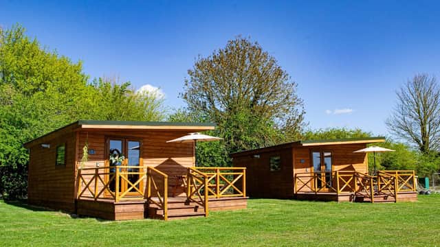 Two wooden cabins with porches on a grassy area, surrounded by trees under a clear blue sky.