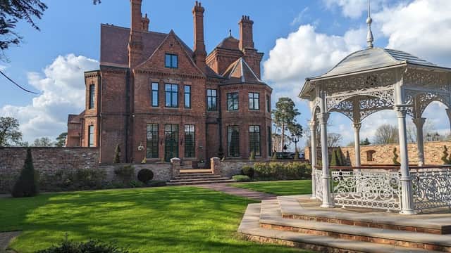 Victorian-style manor with red brick, tall chimneys, and a white gazebo in a landscaped garden under a blue sky.