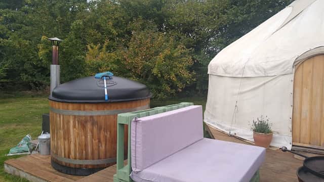 Wooden hot tub with a chimney beside a white yurt and a green bench on a wooden deck, surrounded by trees.