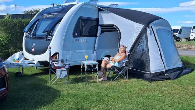 A man relaxes outside a caravan with a sunshade on green grass under a clear blue sky at Melgoats CL Site.
