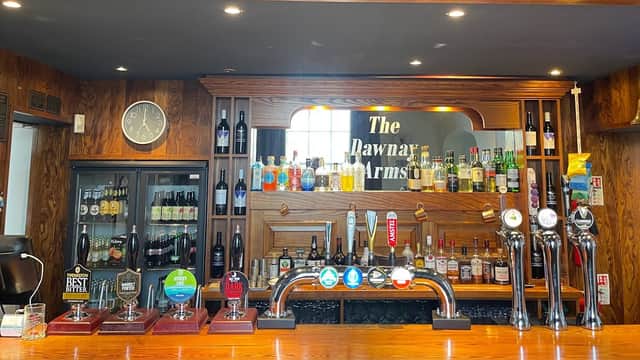 Wooden bar with a variety of beer taps, shelves of bottles, and a clock on the wall at The Dawnay Arms hotel.
