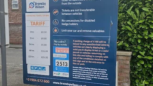 Welcome sign for Carlisle Street Car Park with parking rates and instructions, surrounded by greenery and buildings.