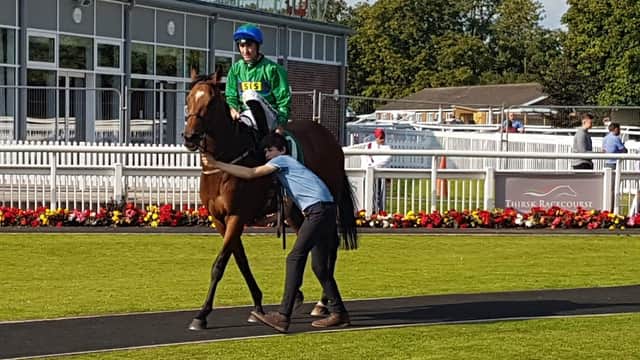 A horse with a jockey in green and blue silks is led by a person along a path at Thirsk Racecourse.
