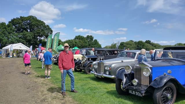 Classic cars lined up in a grassy area with people walking around, tents in the background, under a blue sky.