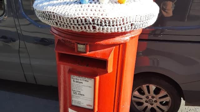 Brightly decorated red post box topped with colorful knitted figures, set against a backdrop of a parked vehicle.