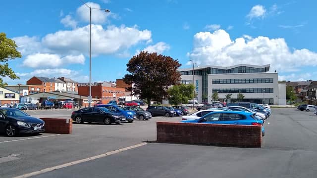 Spacious car park with various parked cars, trees, and a modern building under a blue sky with scattered clouds.