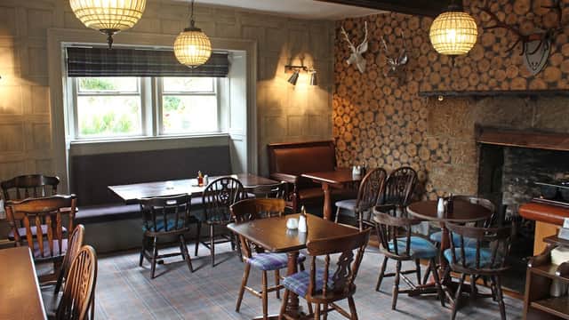 Cozy bar interior with wooden tables, chairs, a stone fireplace, and decorative logs on the wall. Warm lighting.