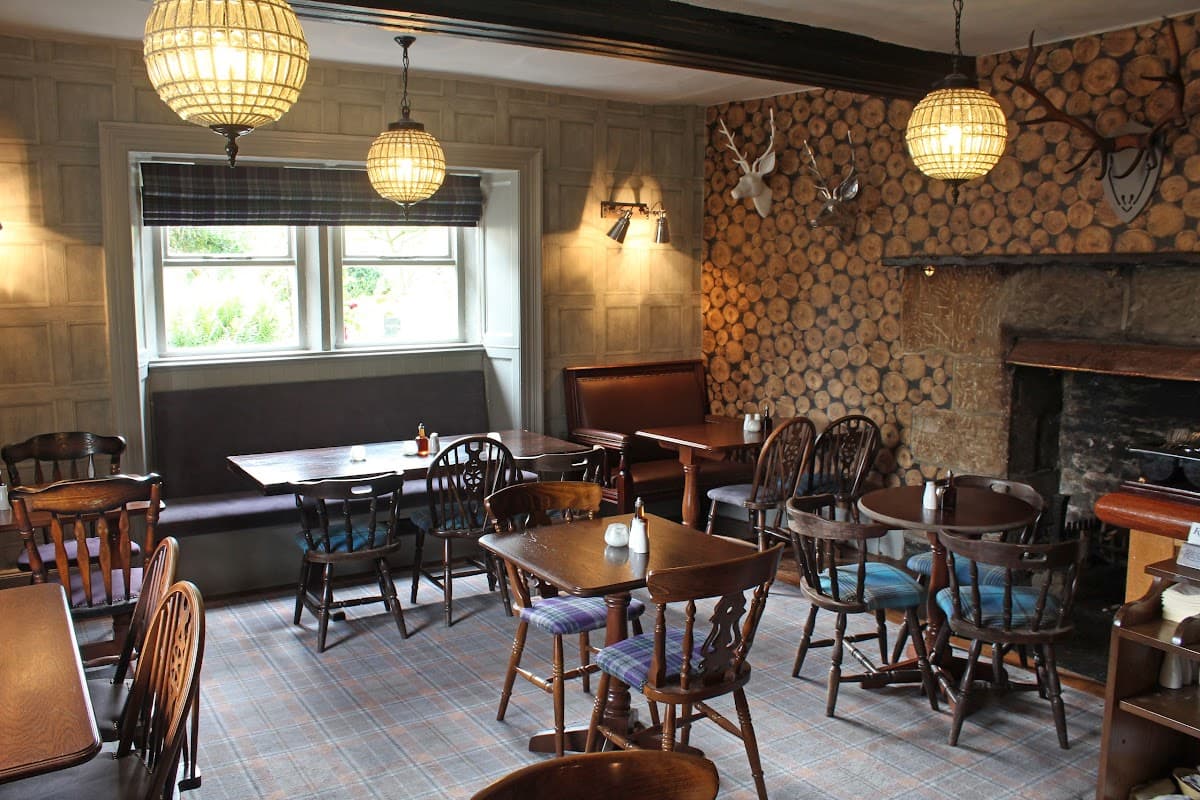 Cozy bar interior with wooden tables, chairs, a stone fireplace, and decorative logs on the wall. Warm lighting.