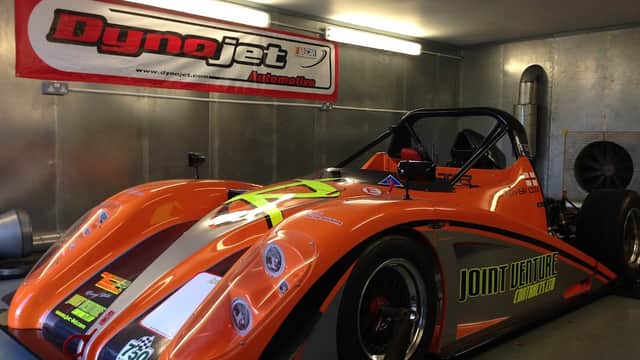 Orange racing car with "Joint Venture" branding inside a garage, featuring a Dynojet banner on the wall.