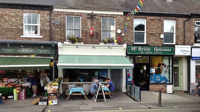 Quaint cafe with outdoor seating, green awning, and colorful bunting, next to a fruit shop and an optician's.