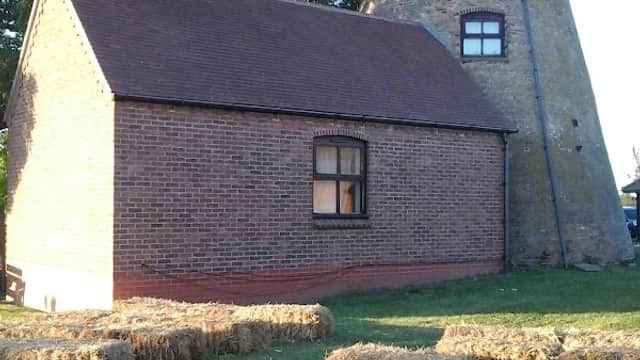 A brick building next to a tall, weathered mill tower, surrounded by straw bales and green grass.