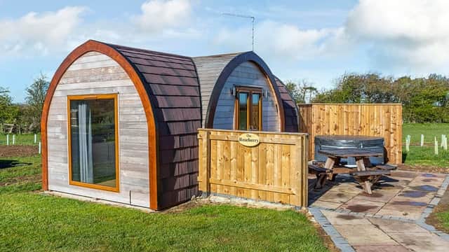 Cozy wooden holiday pod with a picnic table and fenced area, set in a green landscape under a blue sky.