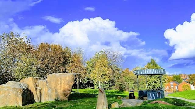 Lush green park with large rocks, trees, and a shelter under a bright blue sky with fluffy white clouds.