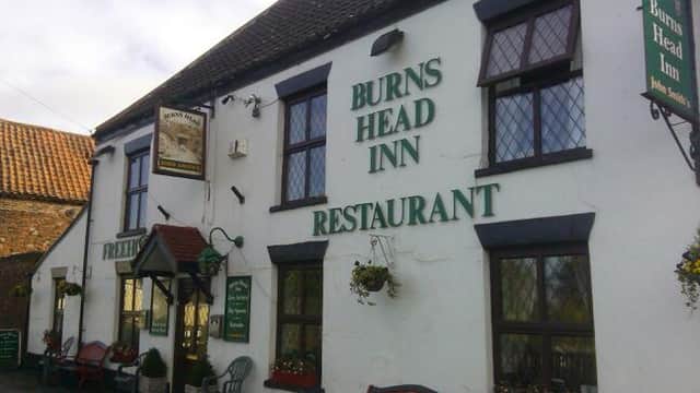 Traditional pub exterior with "Burns Head Inn" sign, green lettering, flower baskets, and benches outside.