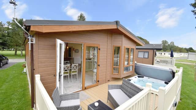 Modern wooden lodge with open doors, patio furniture, and a hot tub, surrounded by green grass and trees.