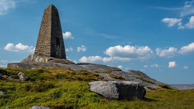 Cracoe War Memorial stands tall on rocky terrain under a blue sky with scattered clouds.
