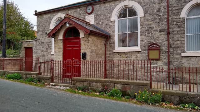Stone building with red door and fence, clock above entrance, surrounded by greenery in Wrelton, Yorkshire.