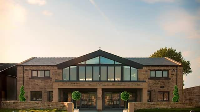 Modern stone building with large glass windows, flanked by manicured greenery and a clear blue sky.