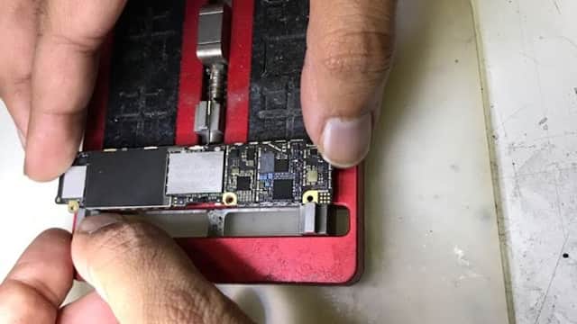 Hands holding a circuit board over a red repair tool, with tools and a work surface visible in the background.