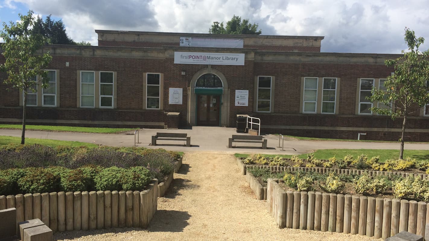 Brick building with large windows, green entrance, and landscaped garden featuring wooden borders and gravel paths.