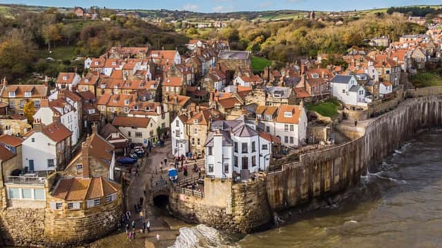 Charming coastal village with red-roofed cottages, rocky shoreline, and a scenic backdrop of hills and blue sky.