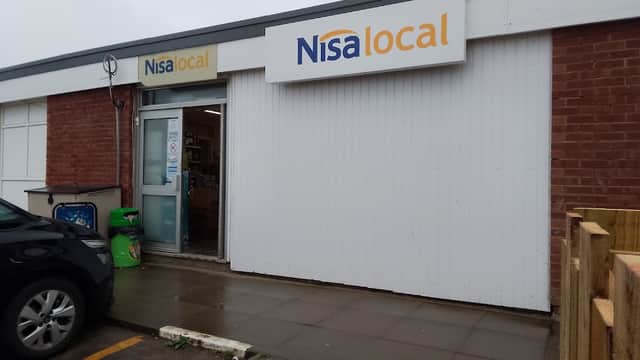 Nisa Local grocery store entrance with a sign, brick wall, and a parked car nearby.