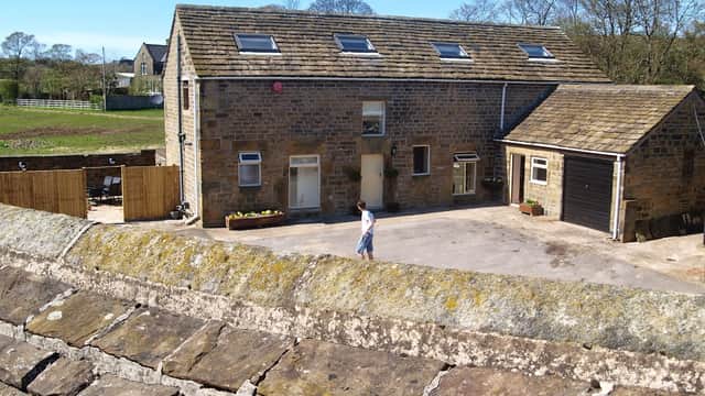 Stone barn with a slate roof, surrounded by greenery, featuring a driveway and a person walking nearby.