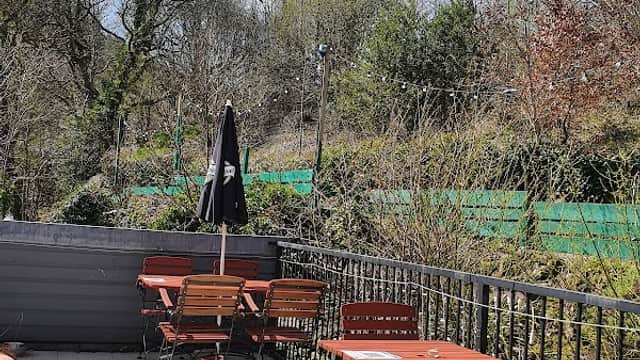 Outdoor seating area with wooden tables and chairs, surrounded by greenery and a black umbrella, under a clear blue sky.