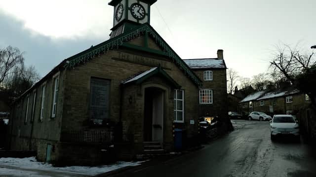 Cononley Village Institute features a clock tower, stone facade, and snowy surroundings with nearby houses on a sloped road.