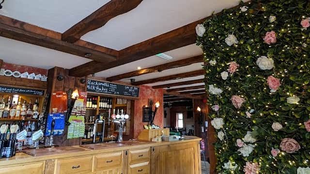 Rustic bar interior with wooden beams, a wooden counter, and a floral wall feature adorned with pink and white flowers.