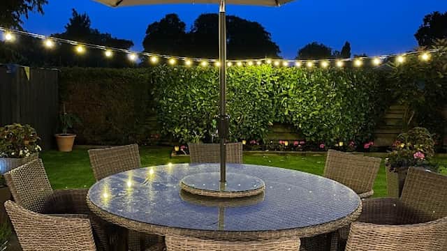 Outdoor dining area with a large round table, wicker chairs, and an umbrella, illuminated by string lights at dusk.