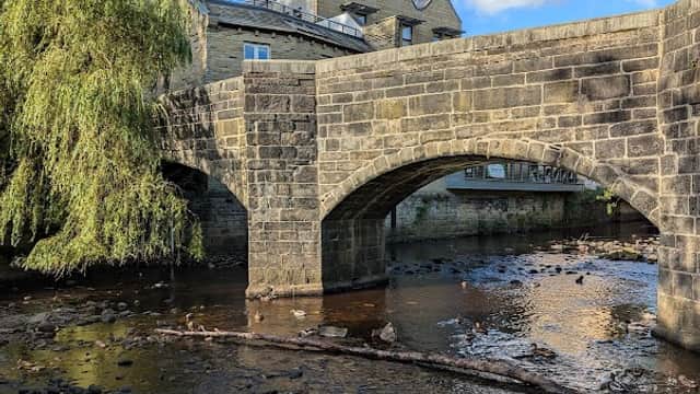 Stone bridge over a river with a weeping willow, surrounded by buildings and blue sky in Chiserley, Yorkshire.