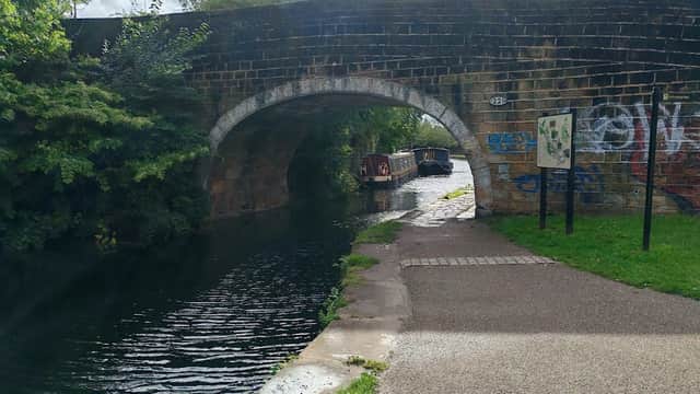 Stone bridge over a canal with moored boats, surrounded by greenery and a pathway beside the water.