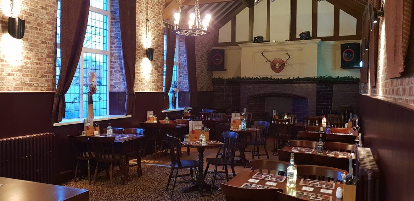 Rustic dining area with wooden tables, chairs, a stone fireplace, and large windows, featuring warm lighting and decor.