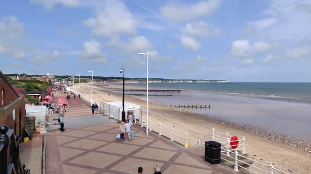 Car park near the beach, with a promenade, sandy shore, and distant cliffs under a partly cloudy sky.